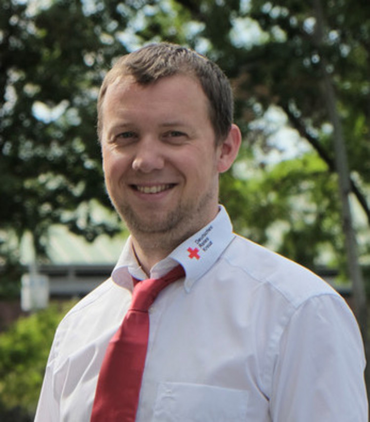 Ein Mann in einem weißen Hemd mit roter Krawatte und schwarzer Hose steht lächelnd auf einem gepflasterten Weg, im Hintergrund sind Bäume.