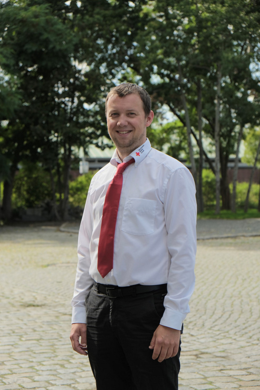 Ein Mann in einem weißen Hemd mit roter Krawatte und schwarzer Hose steht lächelnd auf einem gepflasterten Weg, im Hintergrund sind Bäume.